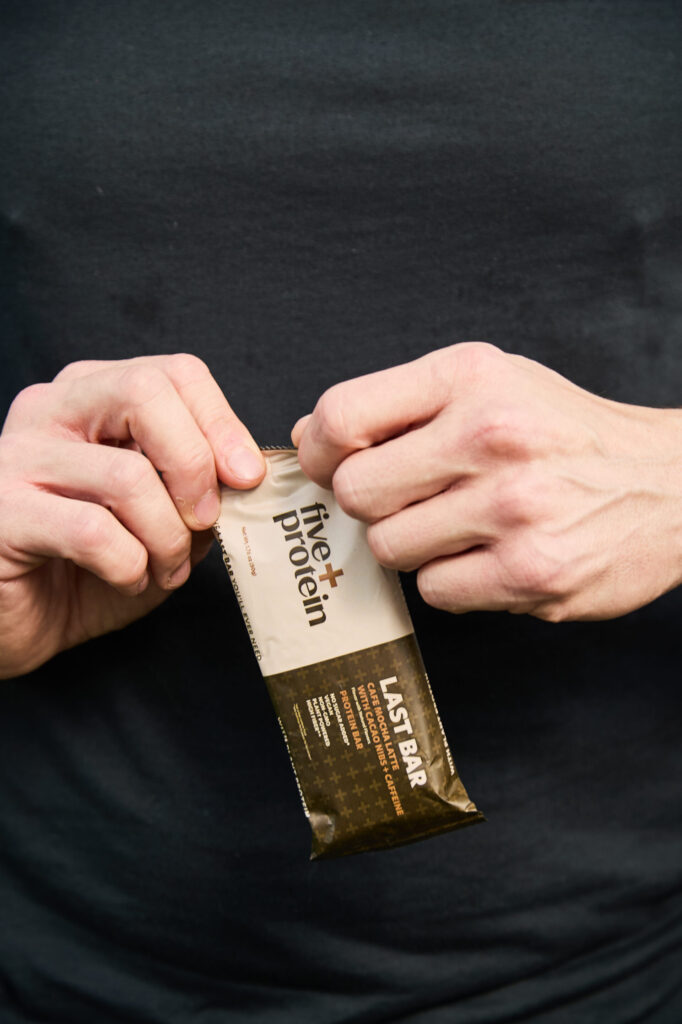 A close-up of a person's hands opening a Five Plus Cafe Mocha Latte protein bar against a black shirt, highlighting convenient high-protein nutrition.