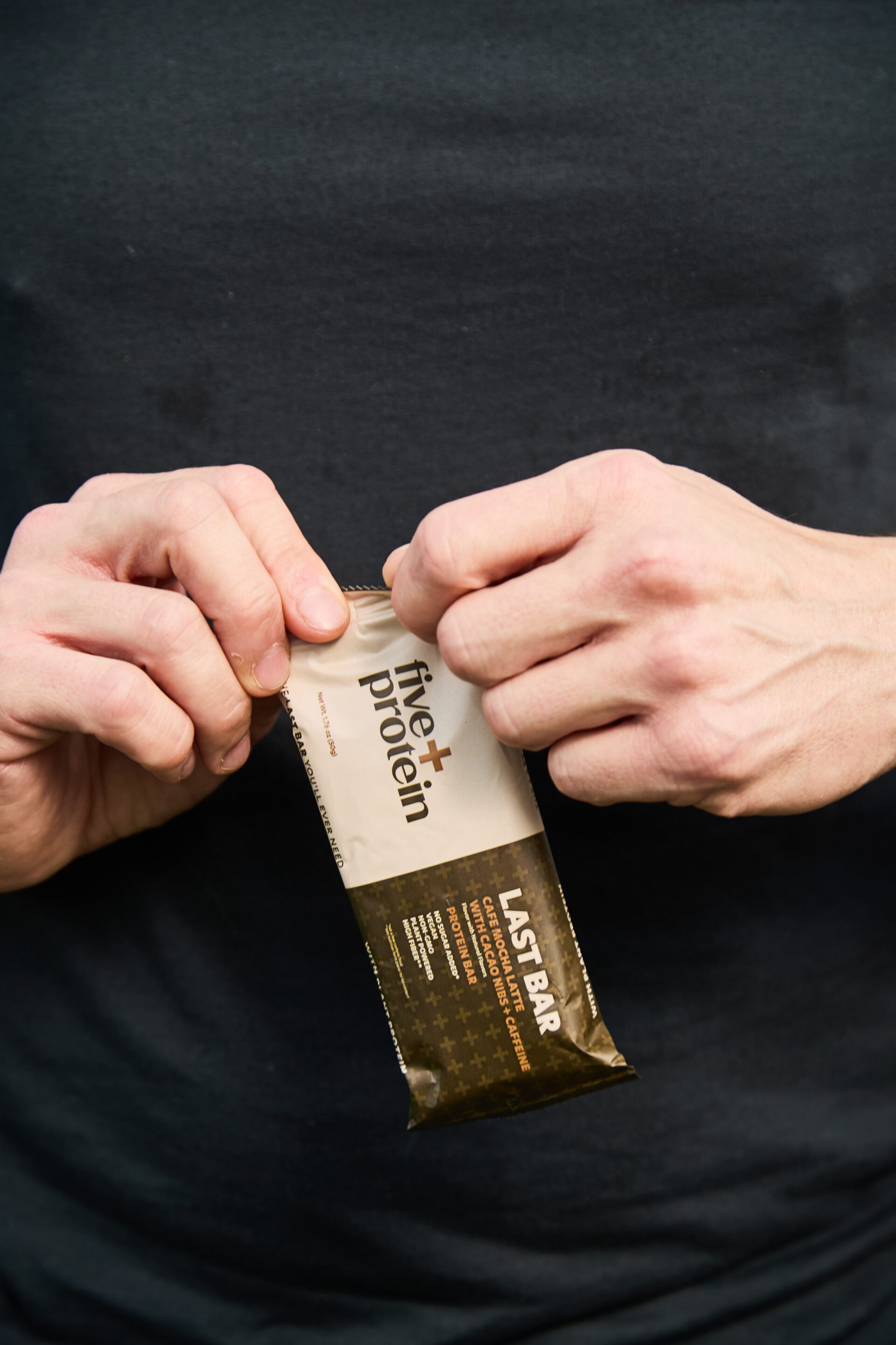 A close-up of a person's hands opening a Five Plus Cafe Mocha Latte protein bar against a black shirt, highlighting convenient high-protein nutrition.