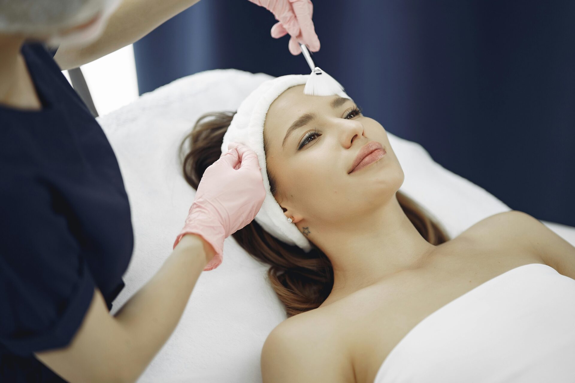 A woman wearing a white headband lies on a treatment table while a skincare professional in pink gloves applies a facial mask with a fan brush to improve her skin health.