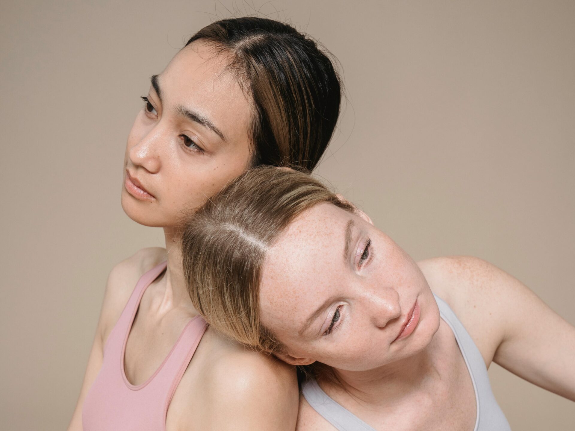 Two women leaning gently against each other with calm expressions against a beige background, representing sisterhood, mutual support, and women's wellness.
