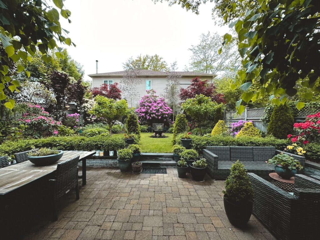 A beautifully landscaped backyard patio with stone pavers, wicker outdoor furniture, potted plants, and a vibrant green lawn lined with blooming purple and red flowers, creating a serene space for mental health.