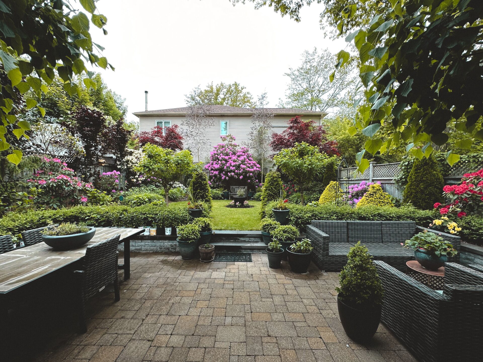 A beautifully landscaped backyard patio with stone pavers, wicker outdoor furniture, potted plants, and a vibrant green lawn lined with blooming purple and red flowers, creating a serene space for mental health.