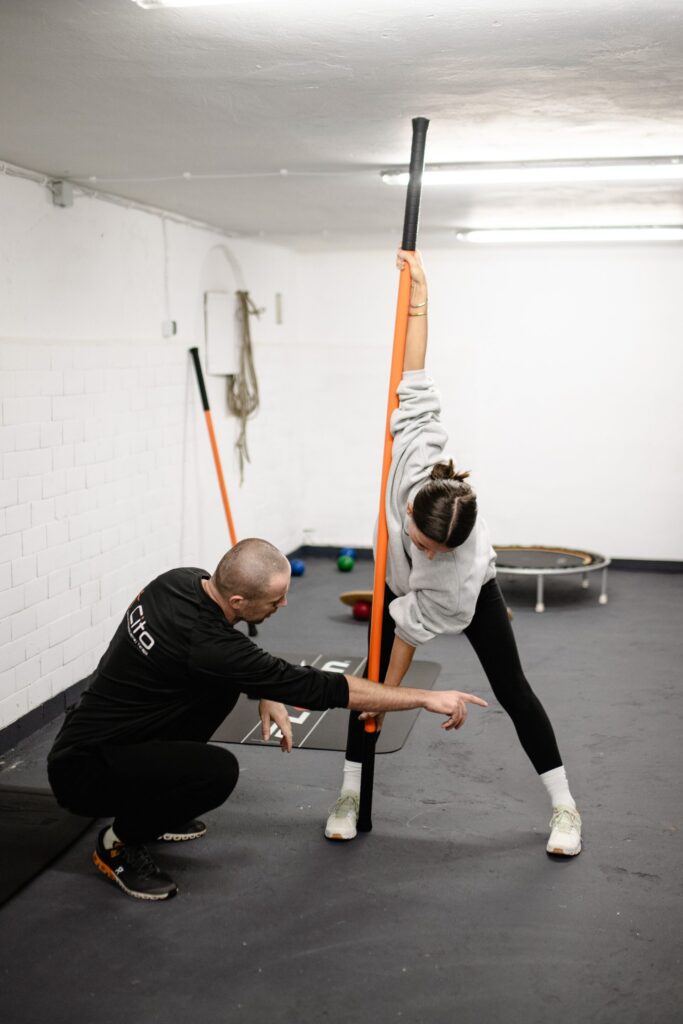 A woman in a grey sweatshirt and black leggings uses a long orange and black training stick to perform a deep side stretch while a male coach guides her form, emphasizing the benefits of mobility training.