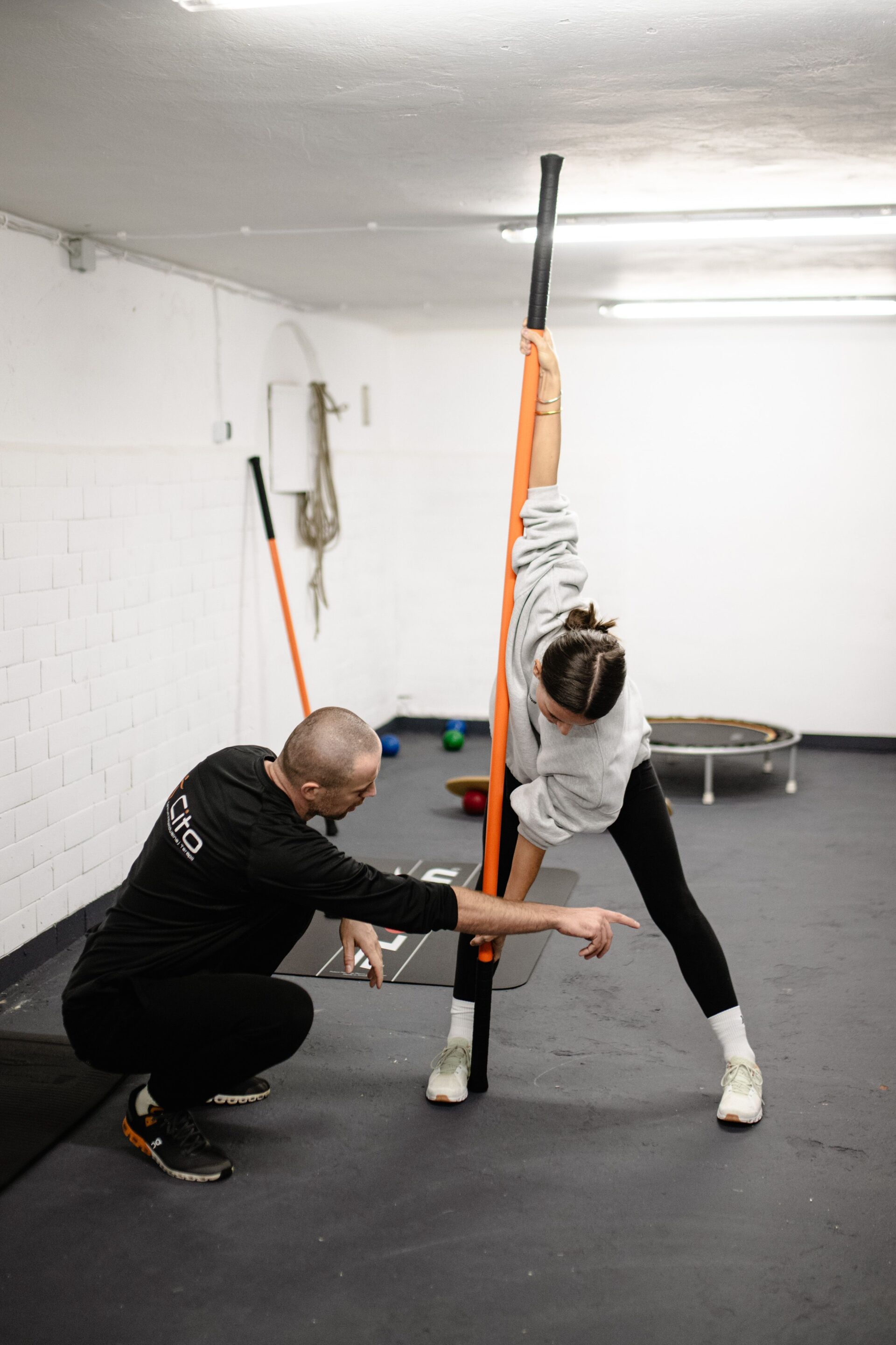 A woman in a grey sweatshirt and black leggings uses a long orange and black training stick to perform a deep side stretch while a male coach guides her form, emphasizing the benefits of mobility training.