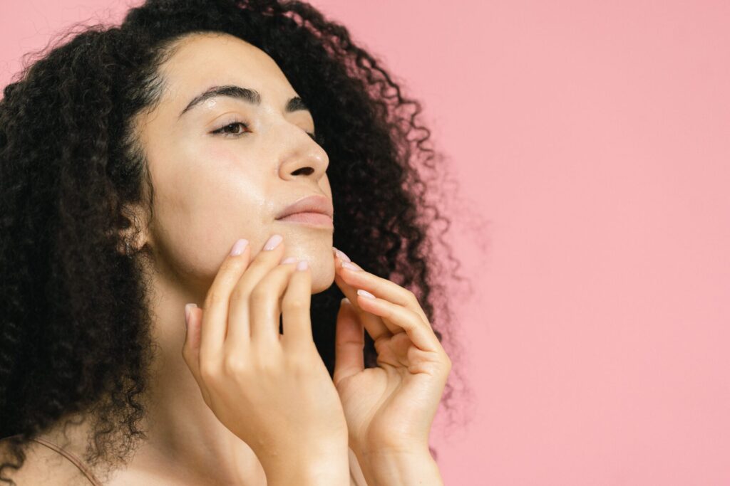 A woman with curly dark hair applying skincare products to achieve a natural skin glow-up against a pink background.