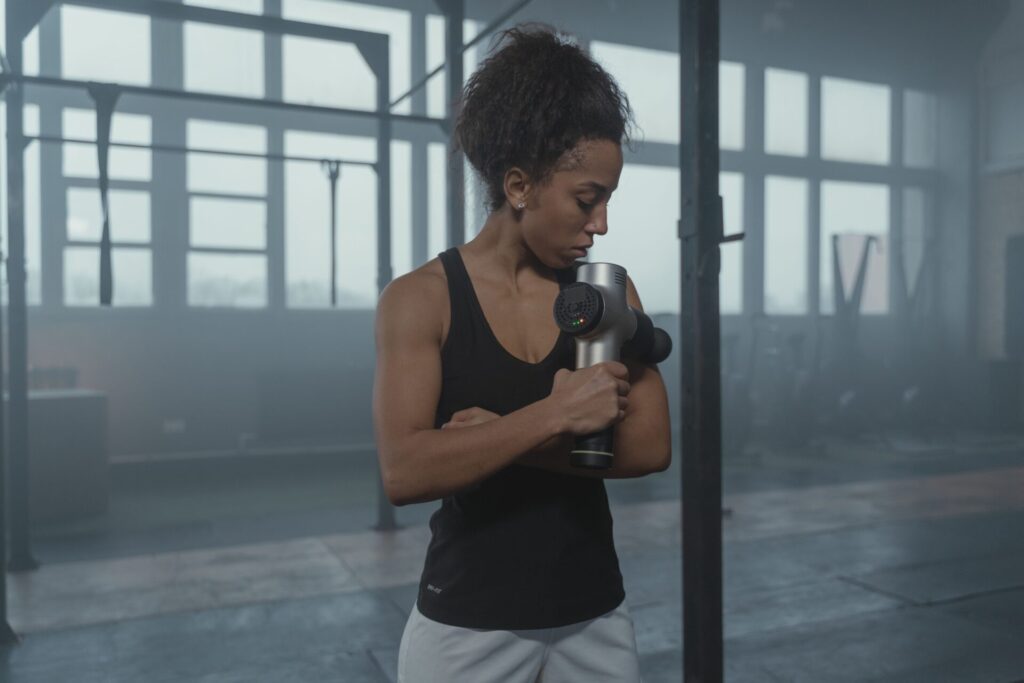 An athletic woman using a massage gun device on her arm for post-workout recovery inside a gym.