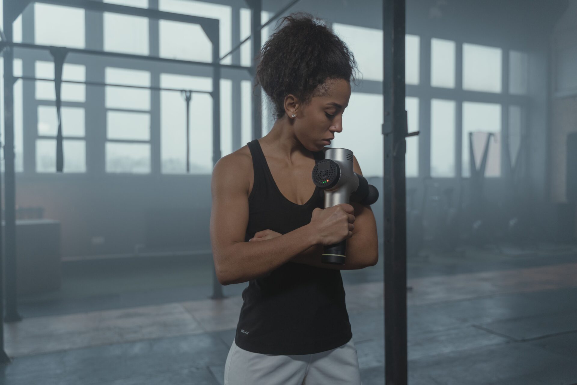 An athletic woman using a massage gun device on her arm for post-workout recovery inside a gym.