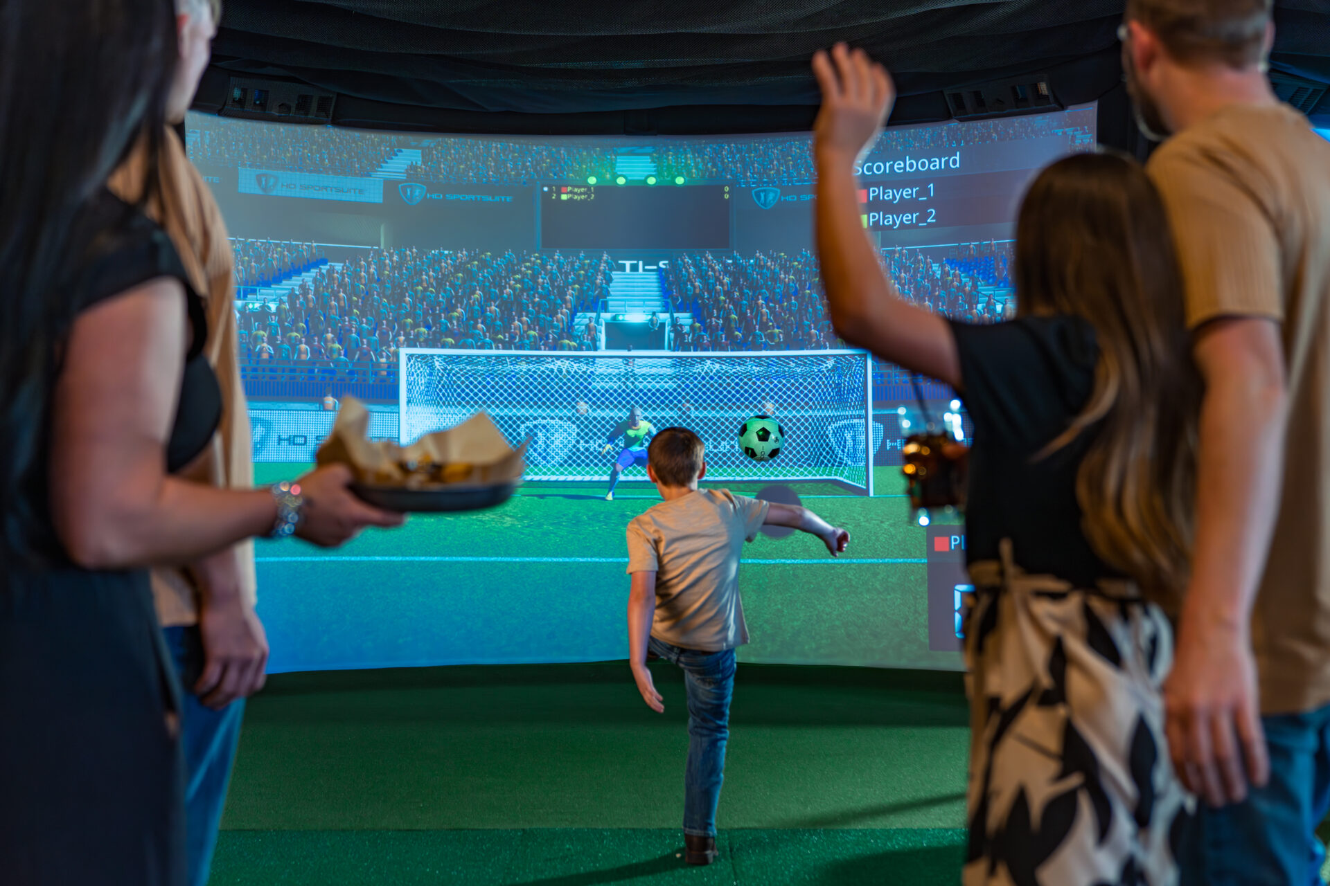 A family watching a young boy kick a soccer ball toward a large virtual screen on an interactive sports simulator at Okana.