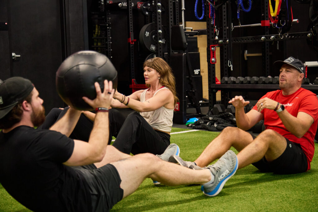 An athlete performing a medicine ball sit-up pass during a guided performance training session on indoor turf.