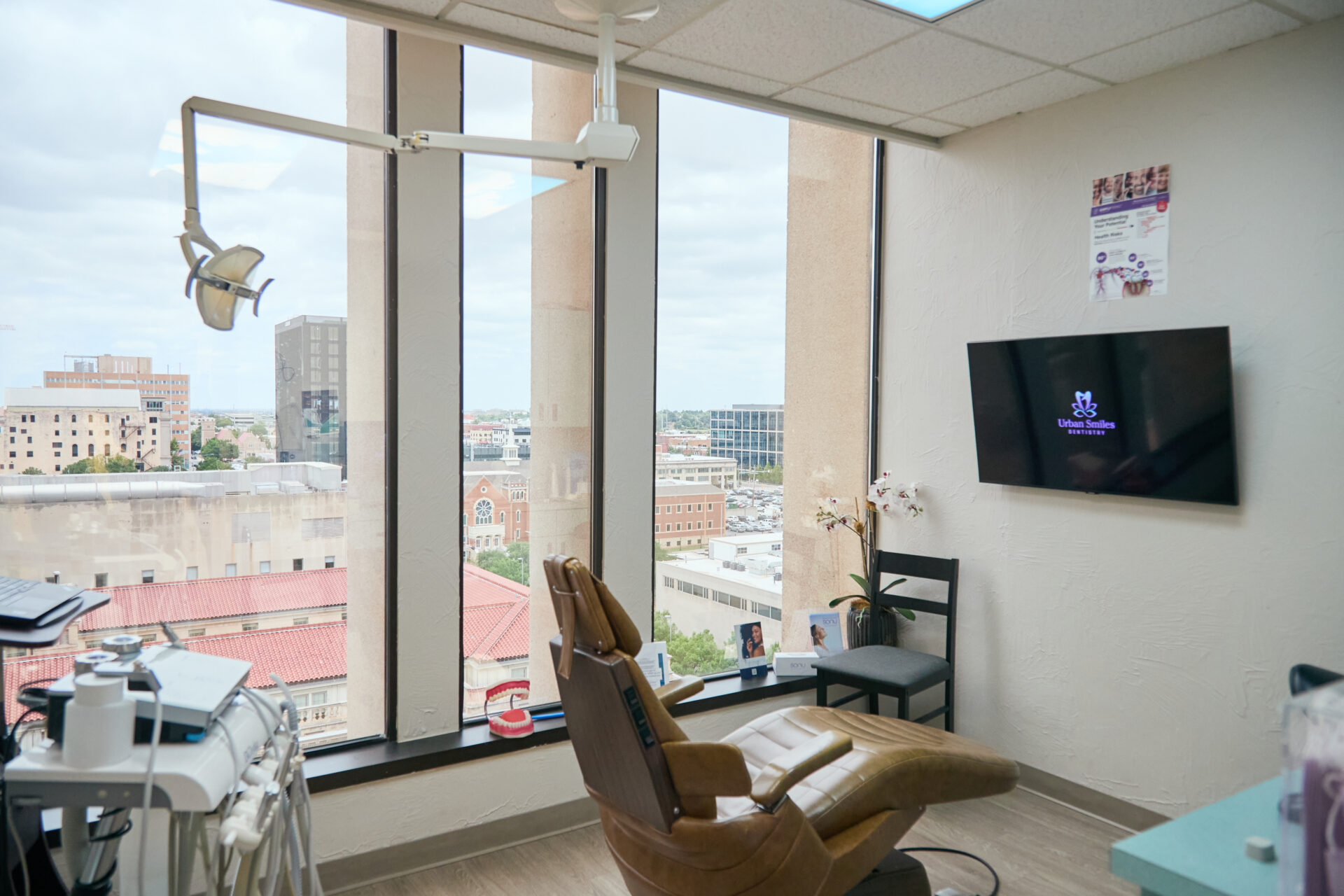 A modern dental treatment room at Urban Smiles Dentistry featuring a leather patient chair, large floor-to-ceiling windows with a city skyline view, and a wall-mounted TV displaying the practice logo.