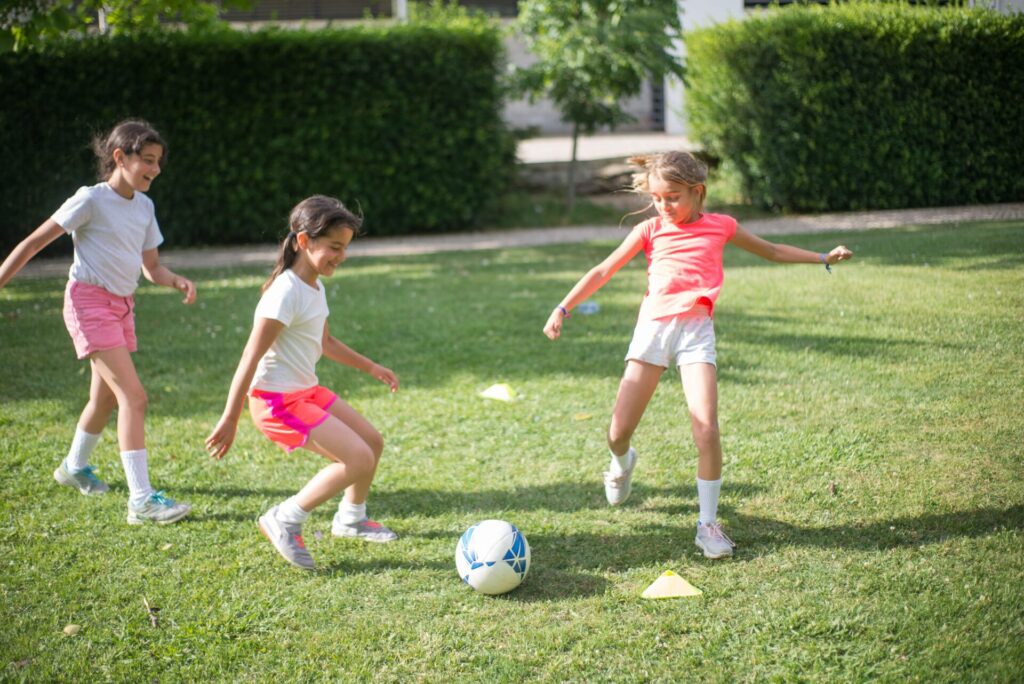 Three young girls playing a game of soccer together on a grassy field during outdoor kids activities.