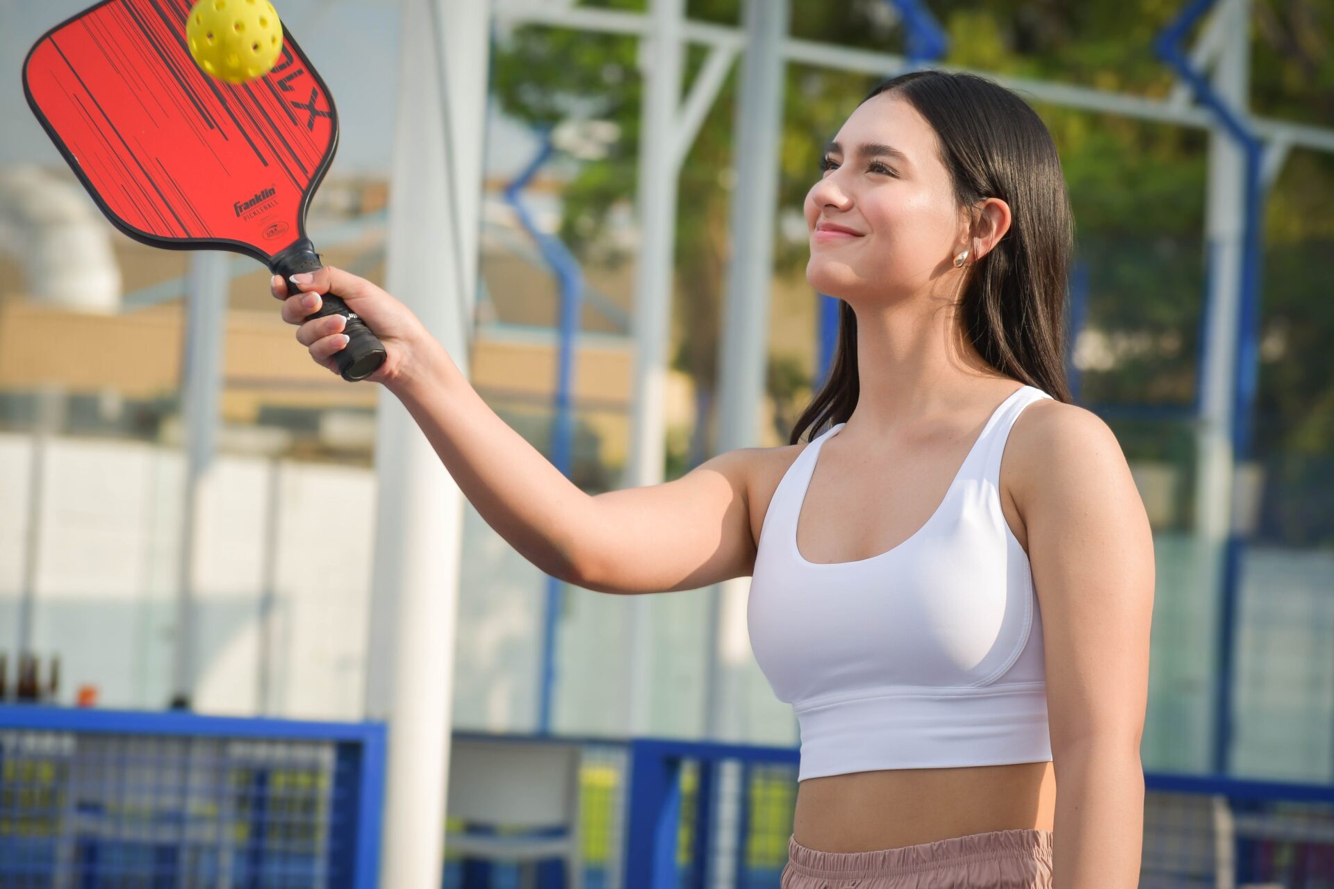 A smiling young woman in a white athletic top holding up a red Franklin pickleball paddle with a yellow pickleball balancing above it on an outdoor court.