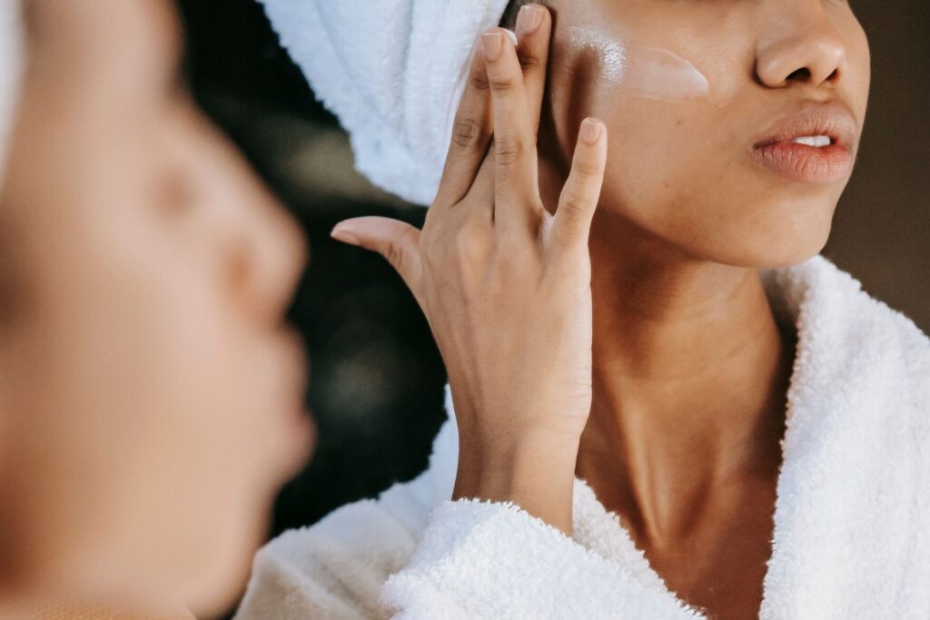A woman wrapped in a white towel and bathrobe applying face cream onto her cheek in front of a bathroom mirror, focusing on skincare from the inside out.