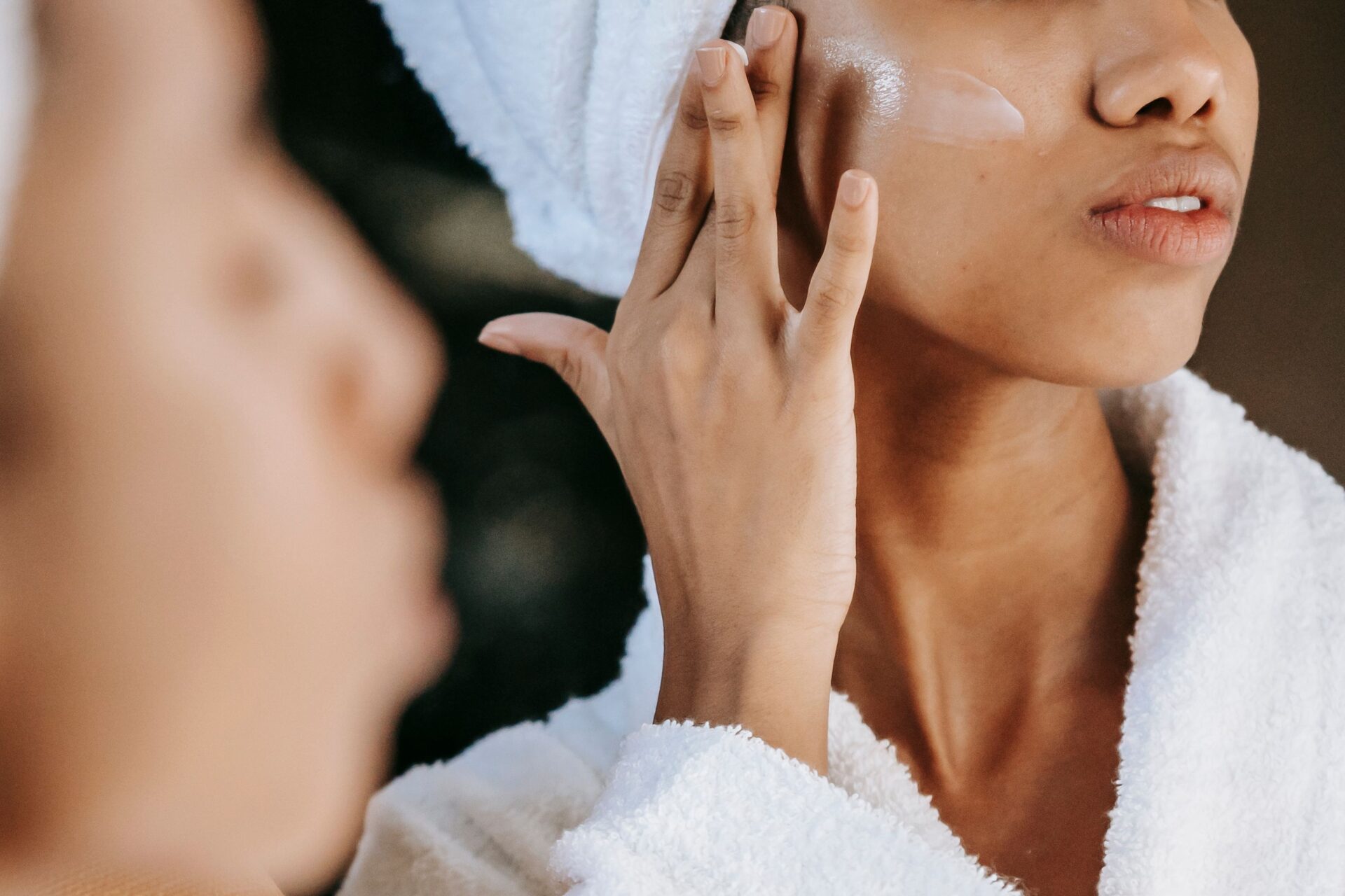 A woman wrapped in a white towel and bathrobe applying face cream onto her cheek in front of a bathroom mirror, focusing on skincare from the inside out.