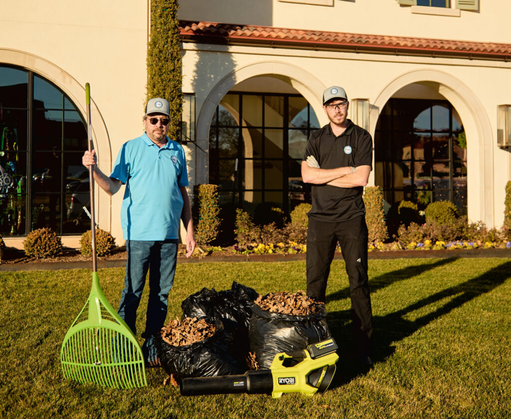 Two Capital City Cleaning team members standing on a manicured lawn outside a large home with a green rake, leaf blower, and bags of gathered yard debris.