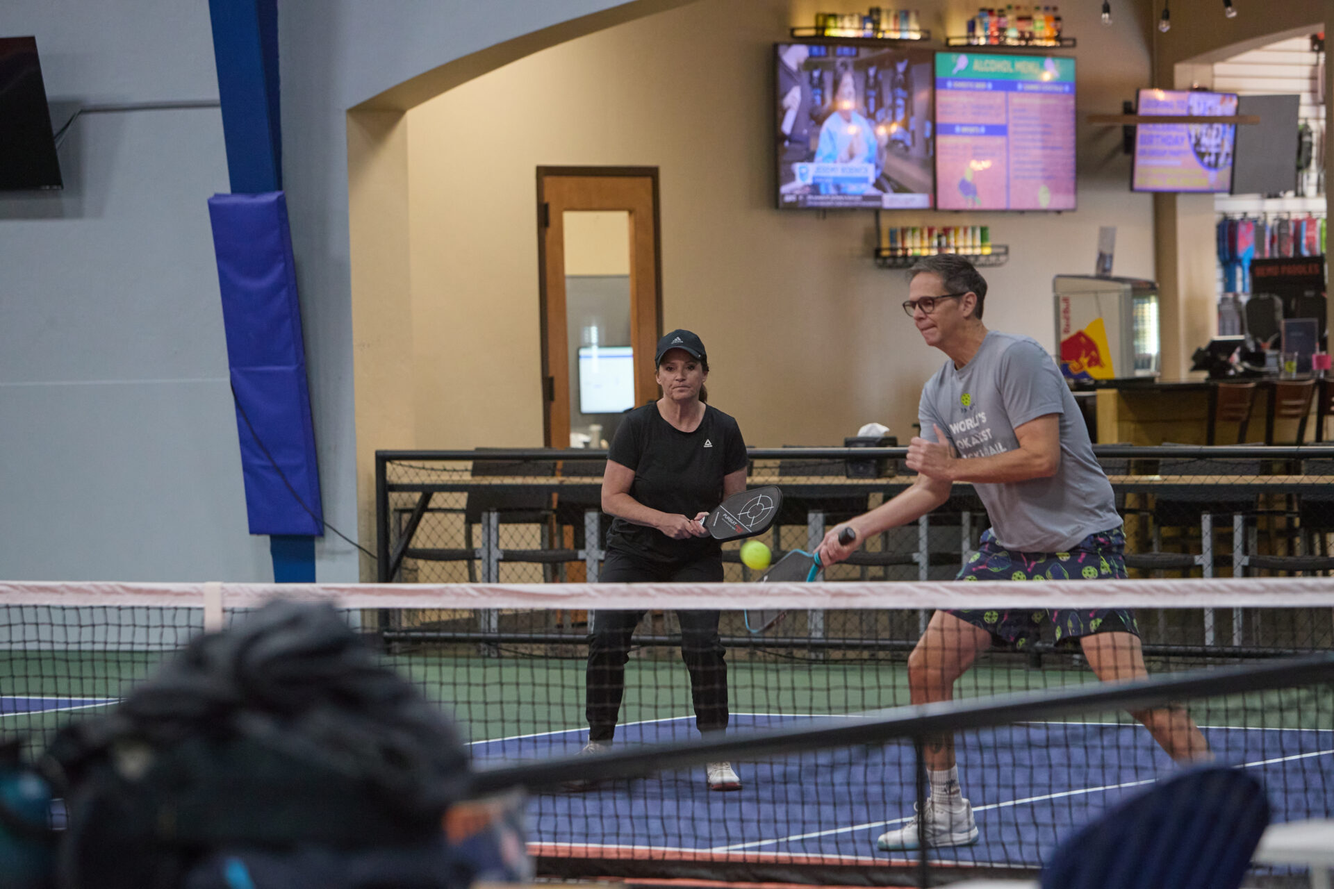 A man and woman playing a doubles match on an indoor court at Hey Pickle Pickle, with the man hitting a yellow pickleball near the net.