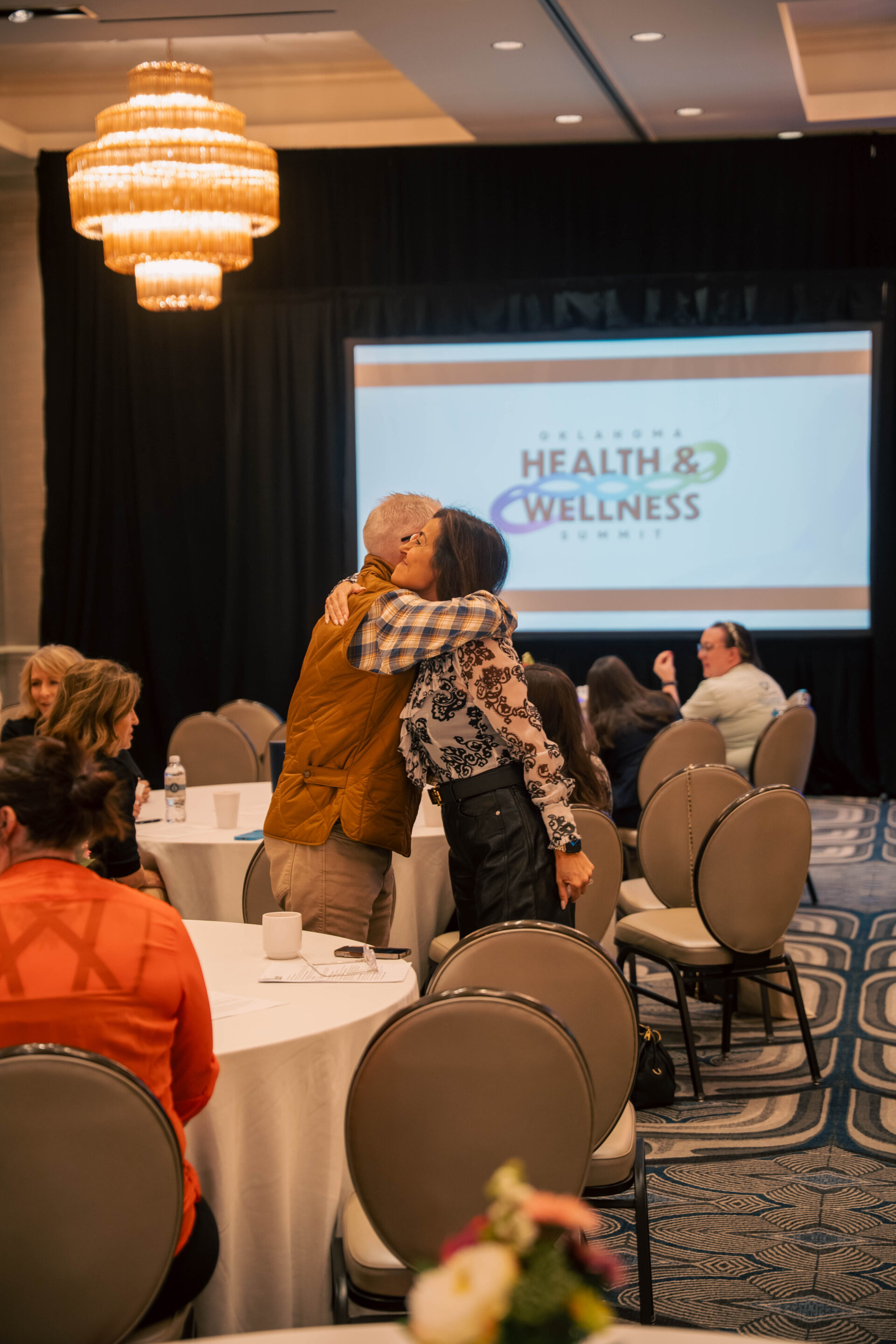 Three male panelists sitting on stage in armchairs during a discussion at The Oklahoma Health and Wellness Summit, with audience members seated at round tables in the foreground.