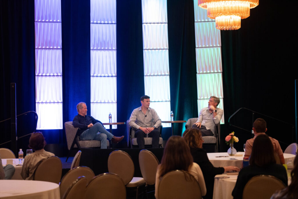 Three male panelists sitting on stage in armchairs during a discussion at The Oklahoma Health and Wellness Summit, with audience members seated at round tables in the foreground.