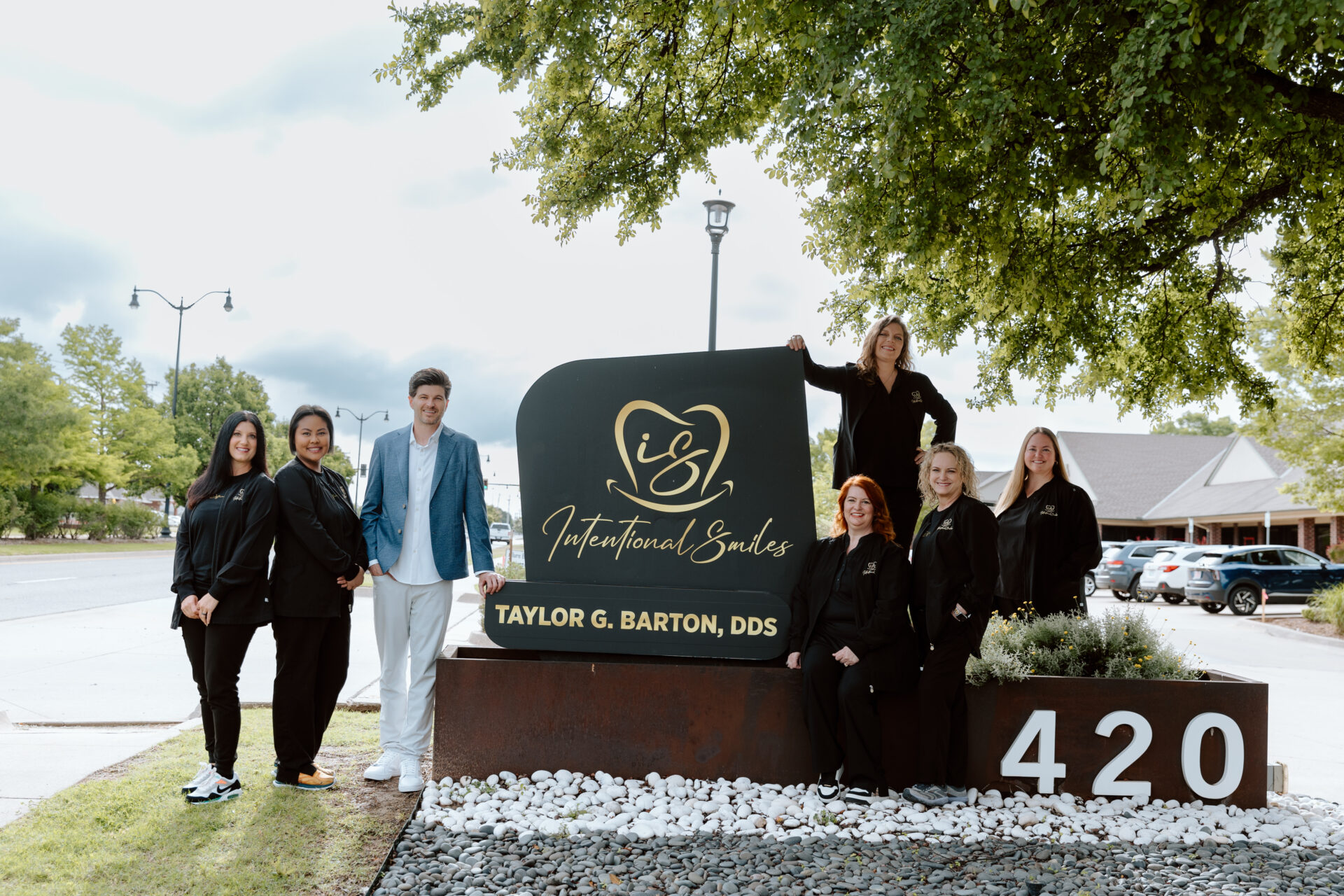 Dr. Taylor G. Barton and the Intentional Smiles dental team posing outdoors next to their dark monument signage with gold lettering and address 420.
